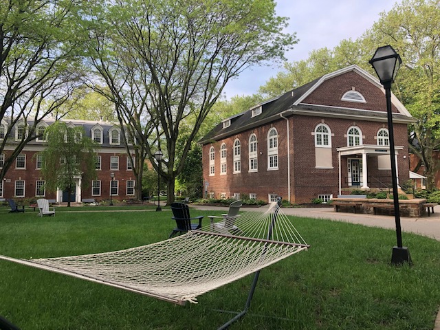 A hammock sits in the sum outside in the quad between 3 campus buildings. A flowering white dogwood tree appear above the roofline of the building behind him. The grass is green.