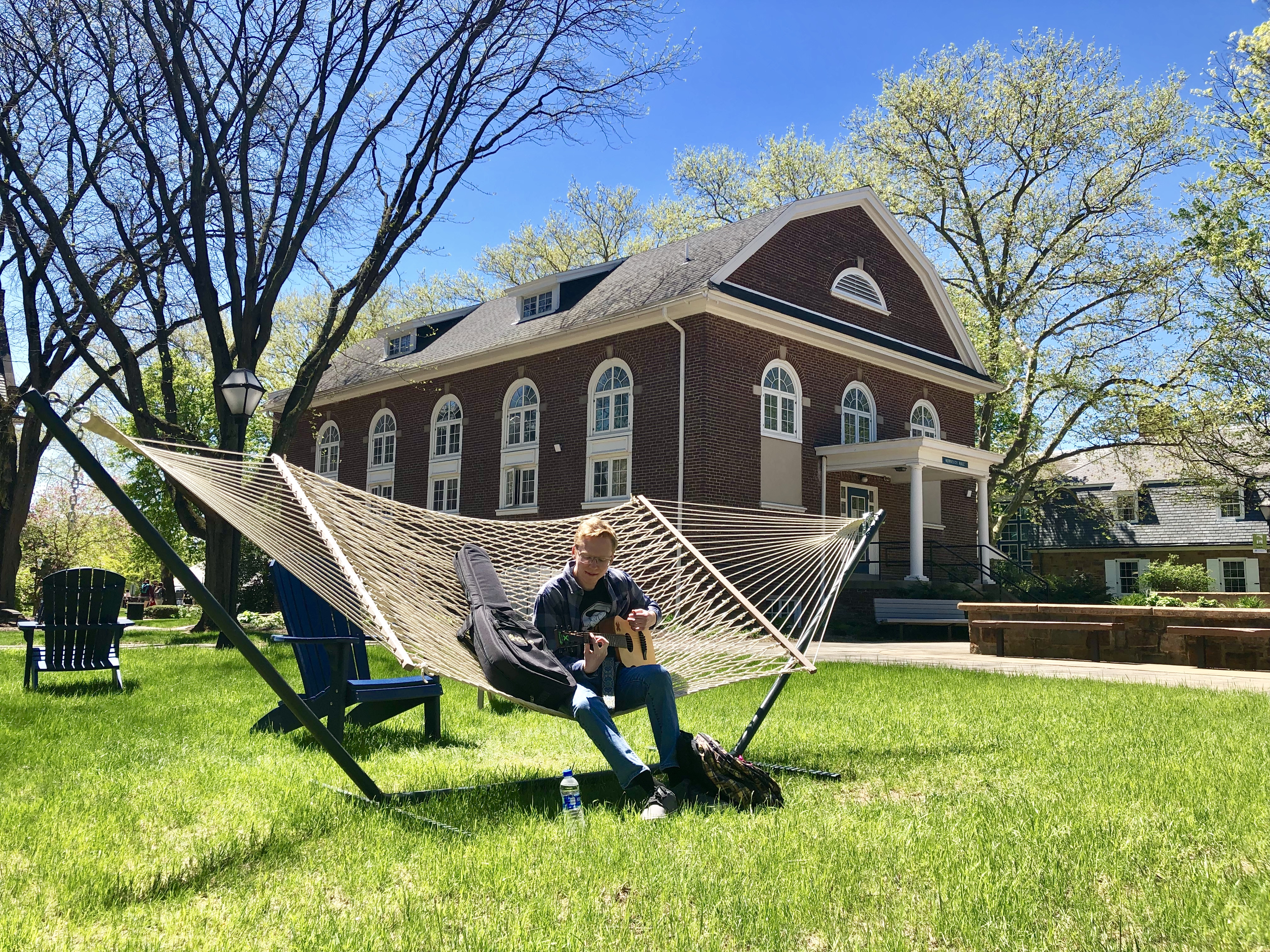 A male student sits on a hammock outside in the quad between 3 campus buildings. A flowering white dogwood tree appear above the roofline of the building behind him. The grass is green.