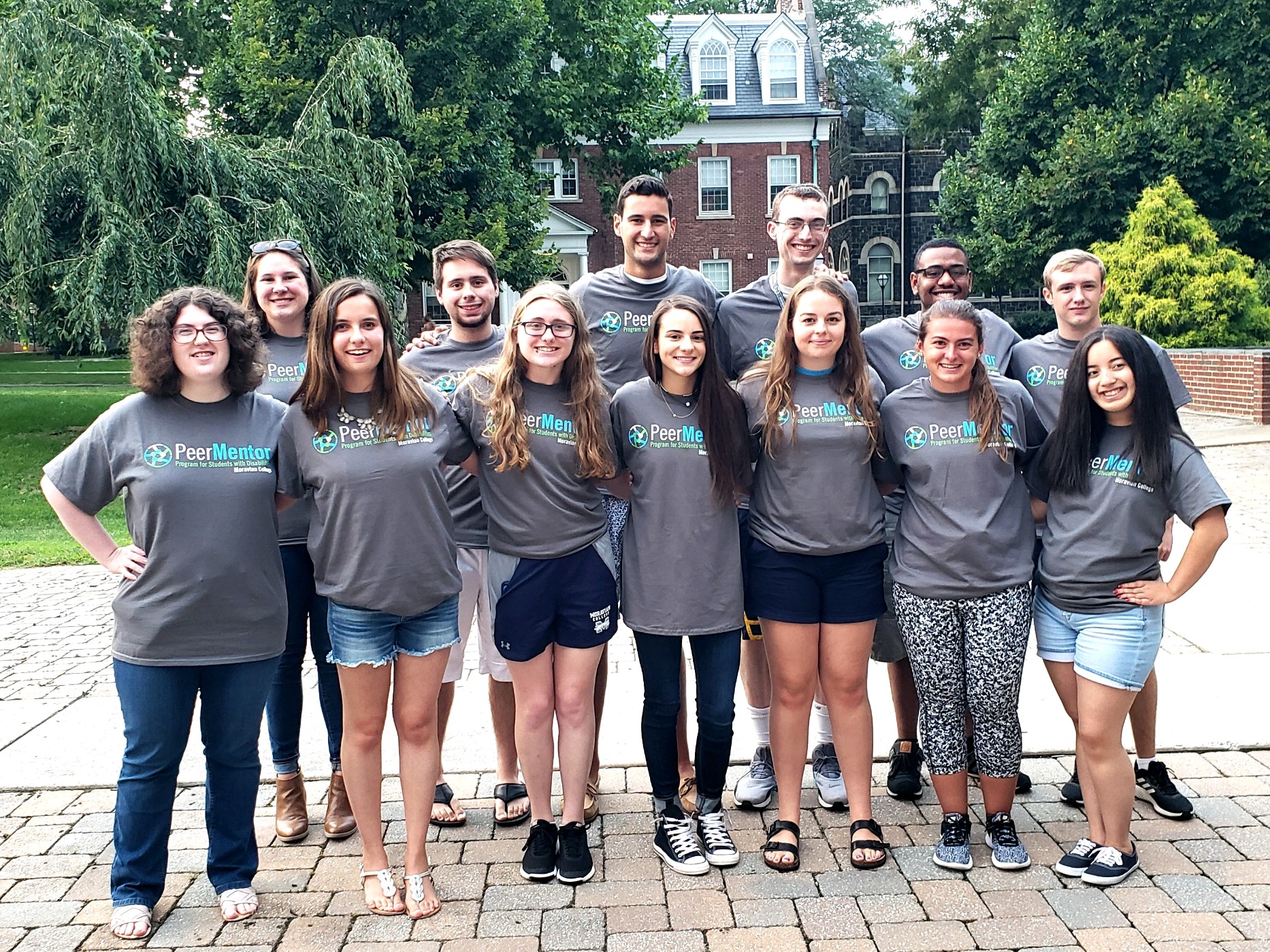 A group of 13 students wearing matching grey t-shirts stand outside posed for a group photo. Their t-shirts read "Peer Mentor Program for Students with Disabilities, Moravian University"