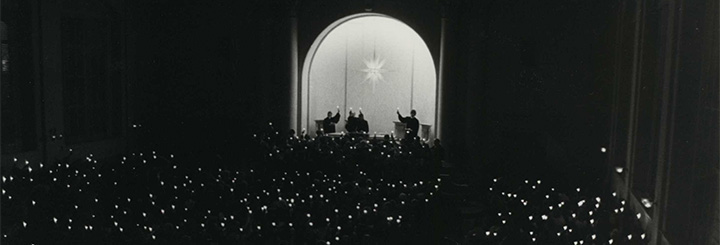 History of the Moravian University Christmas Vespers Banner featuring black and white image of large congregation from behind holding up candles in the dark.