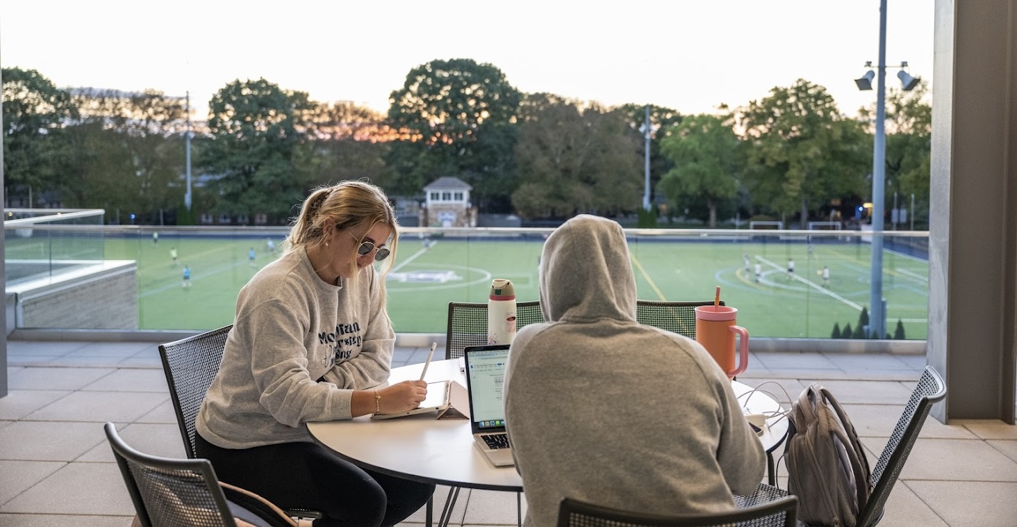 Two students wearing Moravian sweatshirts studying at a table on the HUB Terrace overlooking MAC field