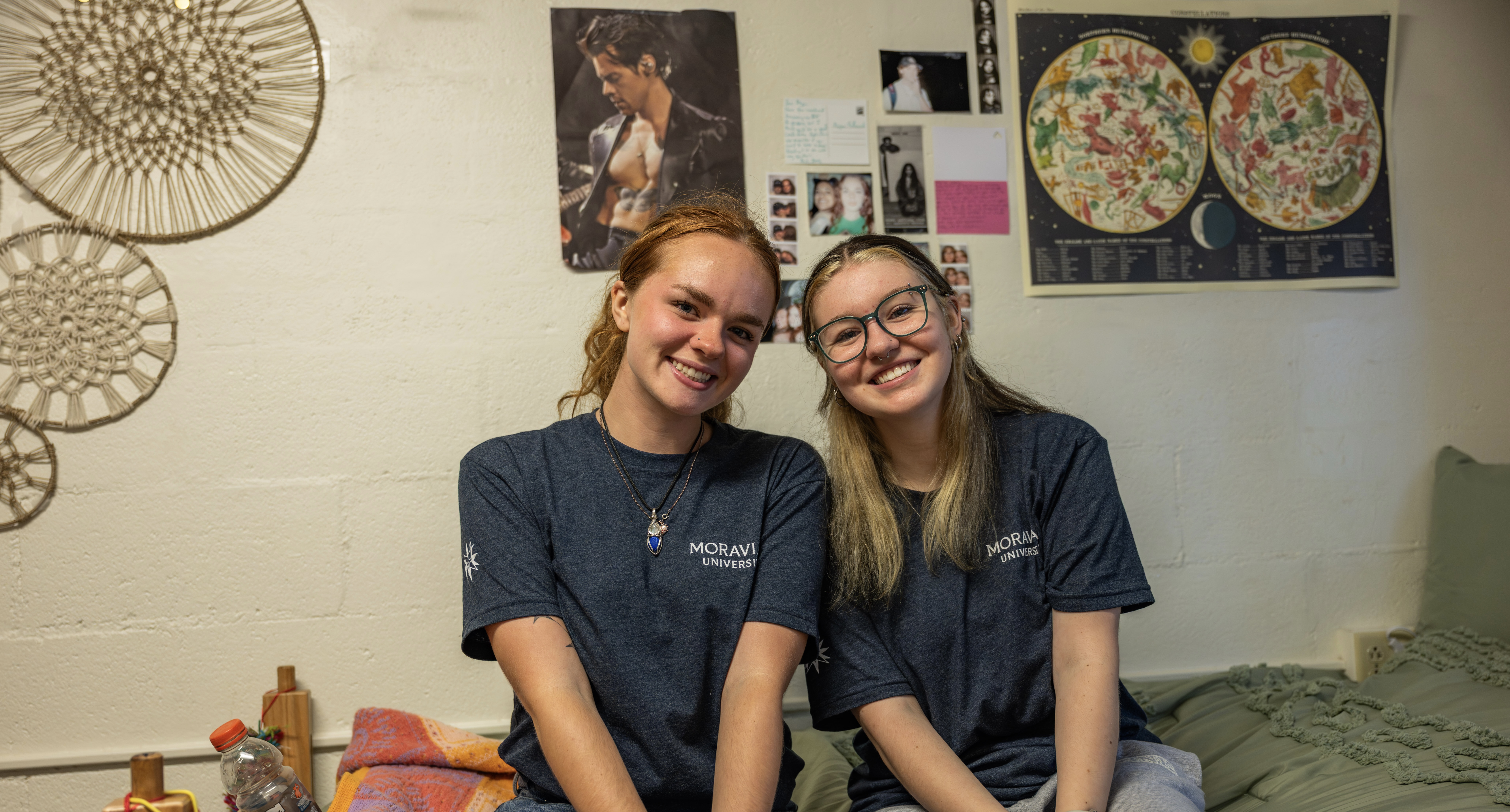 RAs Maggie and Emma in their blue staff t-shirts sitting on a residence hall bed next to each other, smiling at the camera