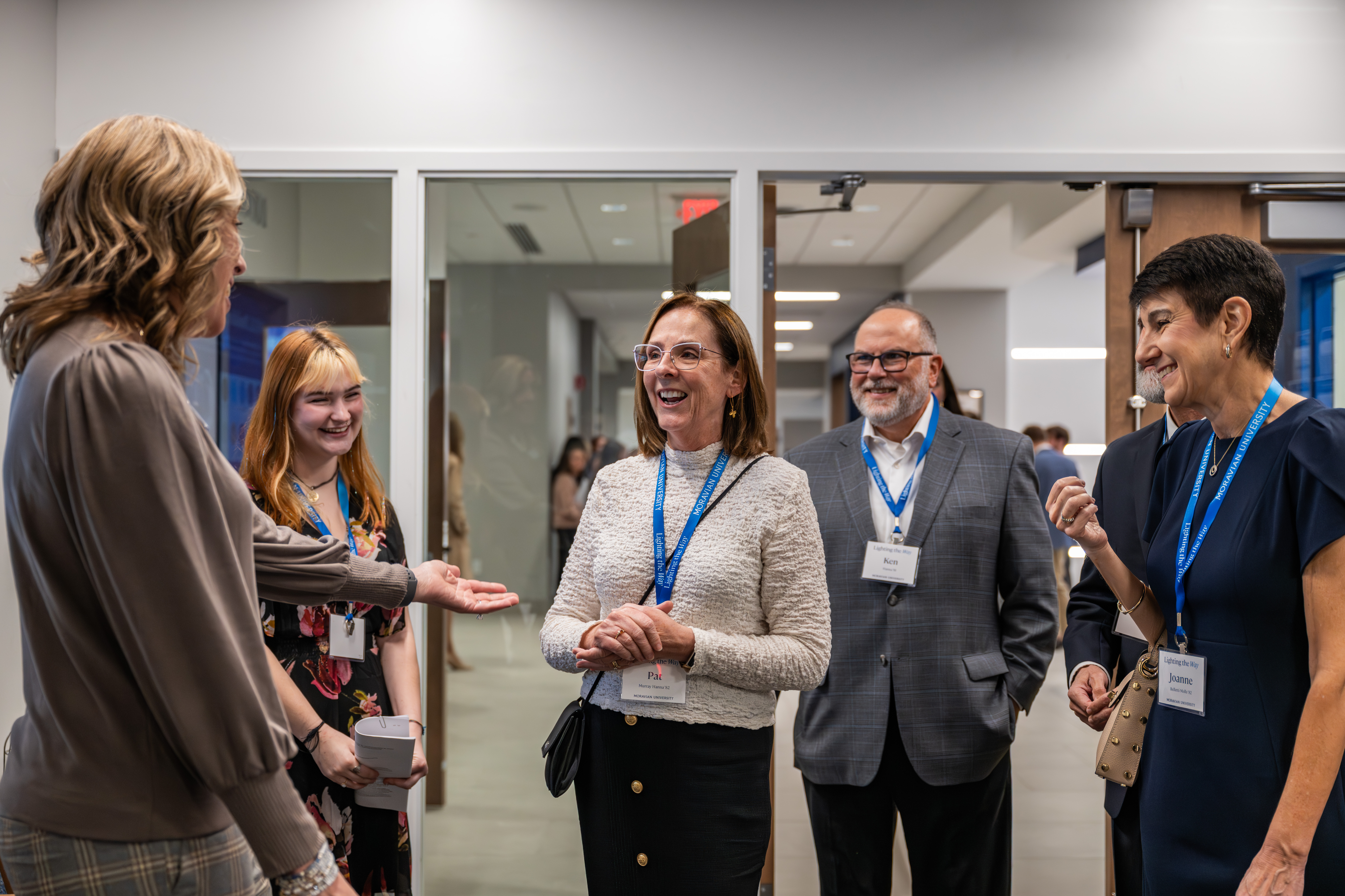 A group of professionals and students engage in a cheerful conversation in a modern hallway. Most participants wear blue Moravian University lanyards and name tags, smiling as they interact. The group includes two women in the foreground speaking with a woman in a white textured top, while a man in a grey suit and another woman look on.
