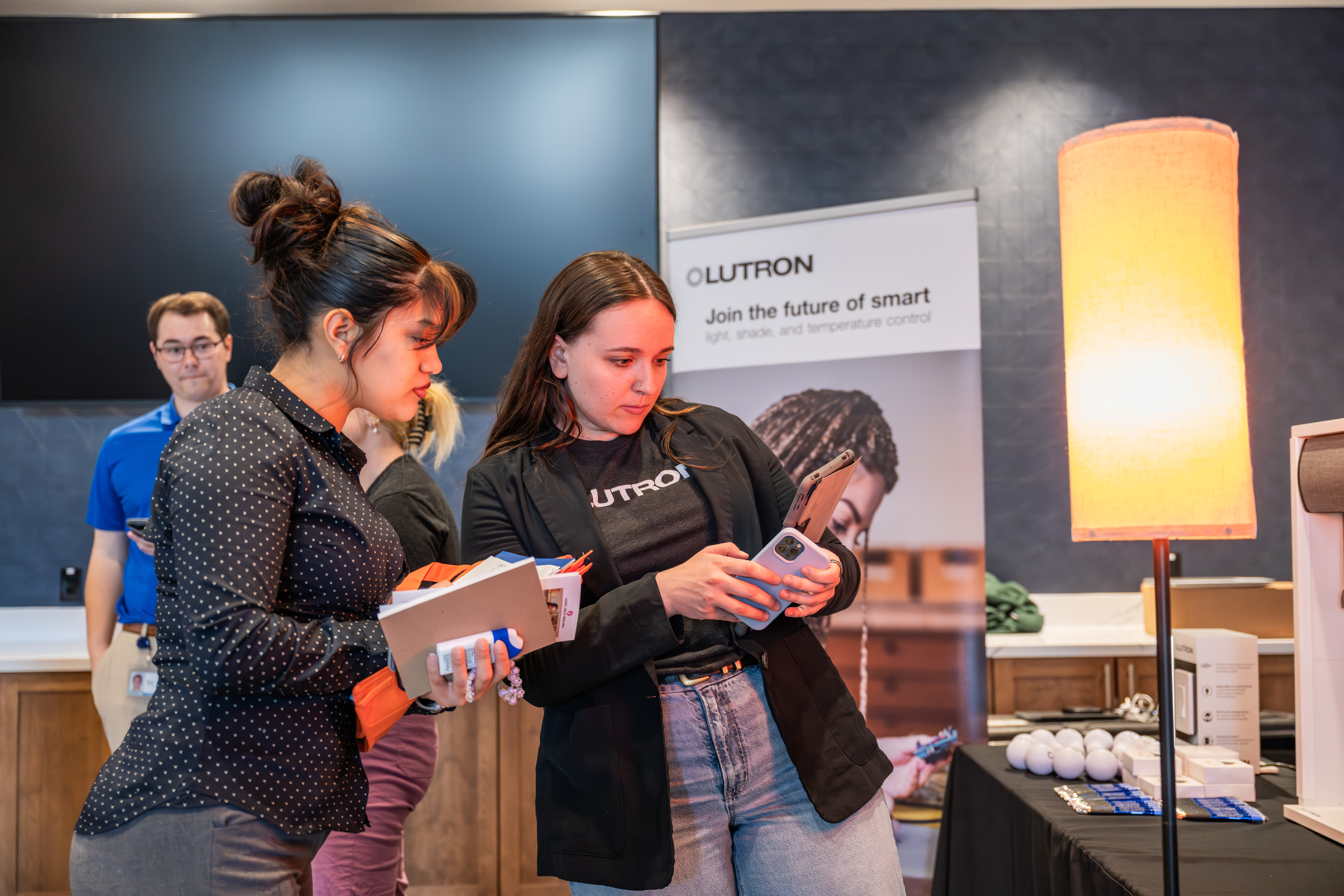 At a career event, a representative from Lutron, wearing a company shirt and black blazer, shows information on her phone to a student holding a notebook. They stand in front of a promotional banner and a table with a glowing lamp, while other attendees are visible in the background.