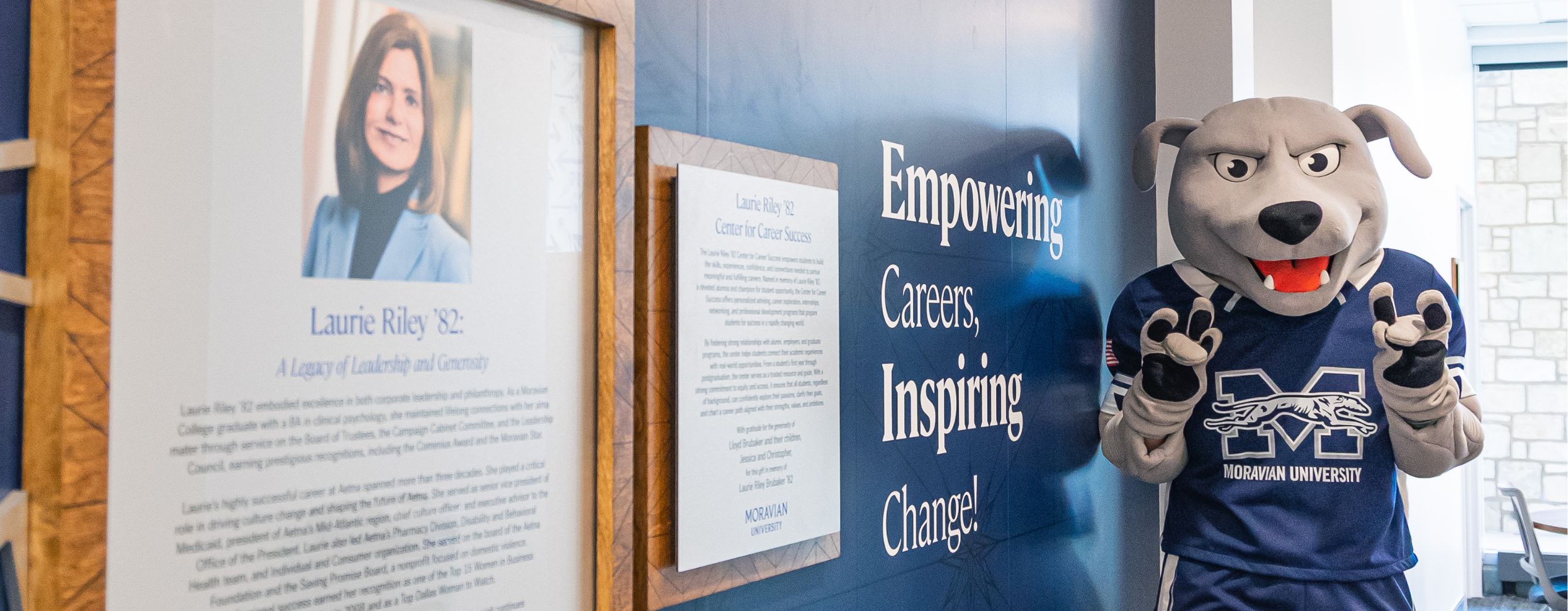 Amos the Greyhound, the Moravian University mascot, poses with paws up next to a commemorative wall for the Laurie Riley ’82 Center for Career Success. The wall features a large framed portrait and biography of Laurie Riley against a navy blue backdrop with the slogan, "Empowering Careers, Inspiring Change!"