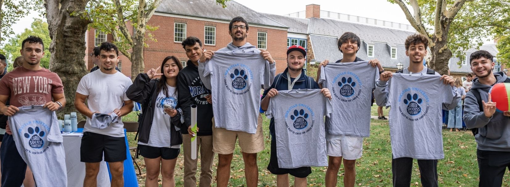  A diverse group of smiling university students standing on a grassy campus lawn, holding up grey T-shirts featuring a blue paw print logo during an outdoor event.
