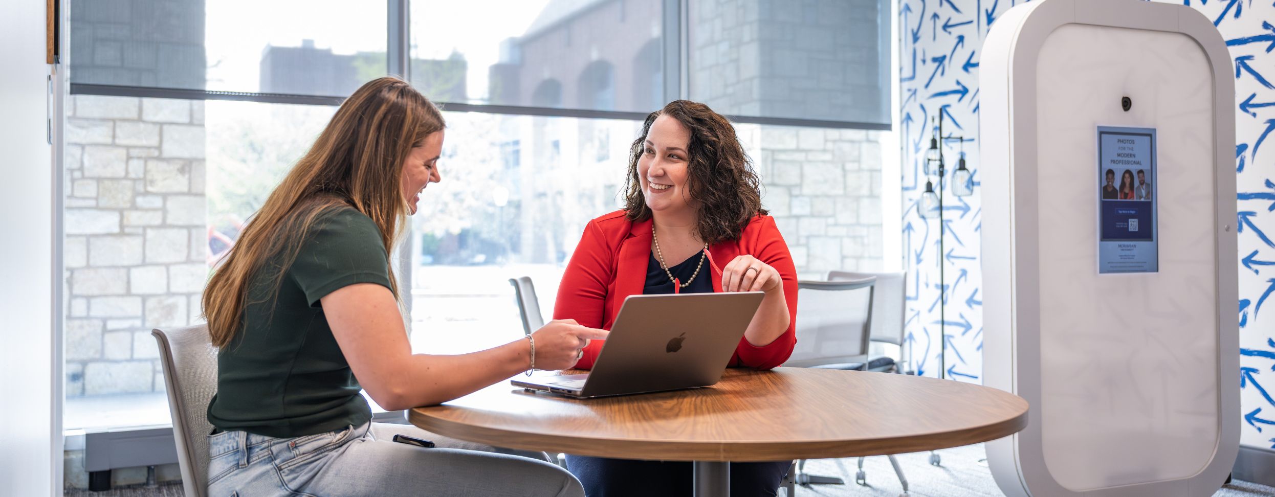Two women sit at a round table in a bright, modern office space, smiling and collaborating over a laptop. Large windows and a professional headshot photo booth are visible in the background.