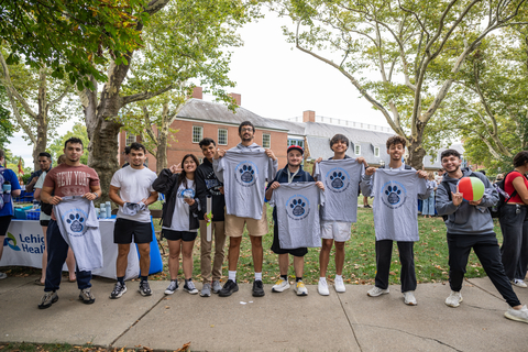  A diverse group of smiling university students standing on a grassy campus lawn, holding up grey T-shirts featuring a blue paw print logo during an outdoor event.