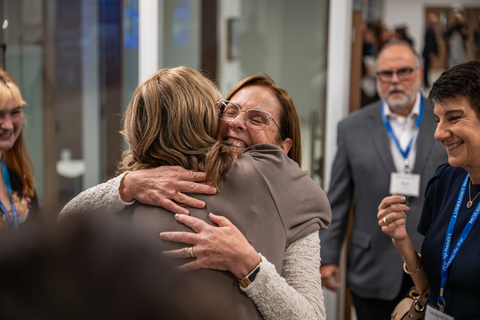 Two women share a joyful, tight embrace at a networking event. One woman faces the camera with a smile and closed eyes, while the other is seen from behind in a white textured cardigan. In the background, other guests in professional attire look on with smiles.